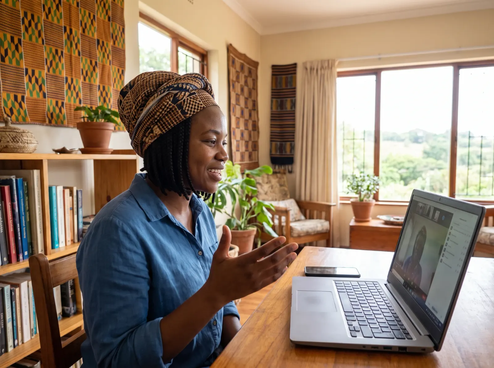 A teacher wearing a patterned head wrap speaks warmly to a student over a video call from her living room
